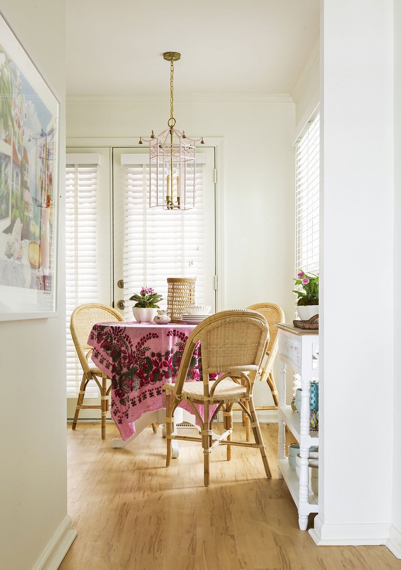Modern boho cottage kitchen table nook with pink and brass chandelier over a white table with hot pink tapestry tablecloth and rattan chairs.