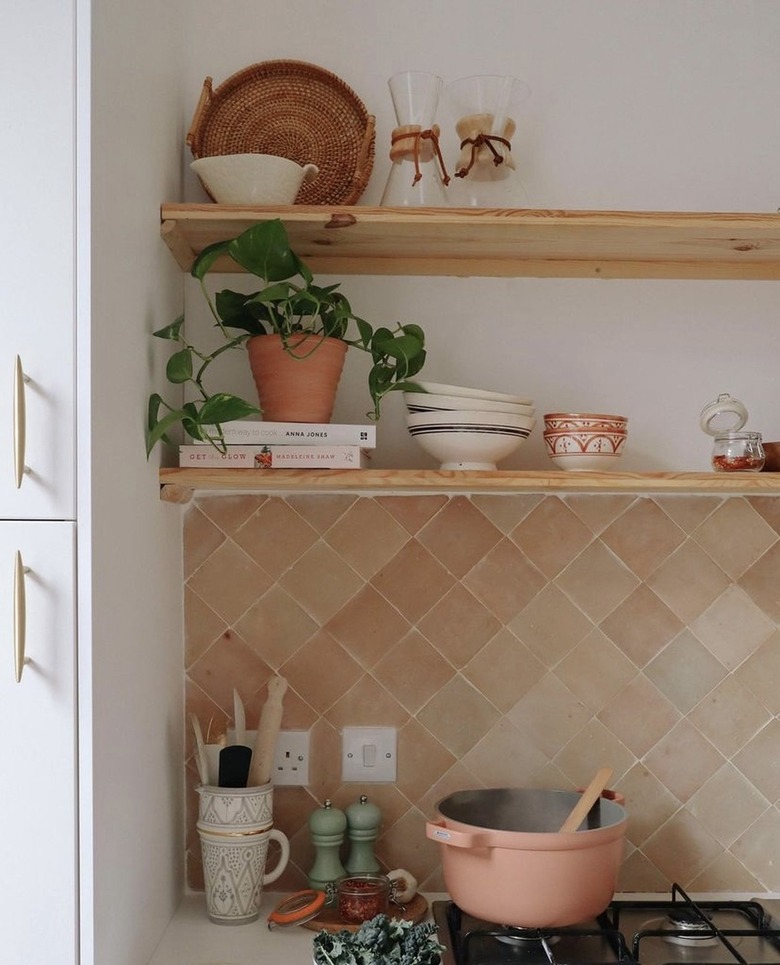 A pink Perfect Pot rests on a stovetop with the lid off and a wooden spoon inside. There is a light pink backsplash and open wood shelving with dishes