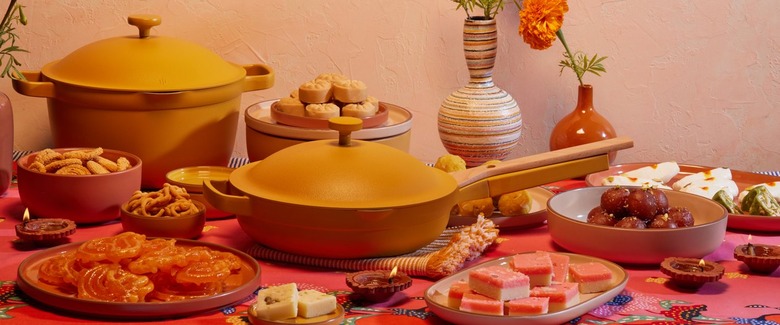 Our Place's turmeric-colored frying pan and pot surrounded by various Indian foods on a table covered in a red cloth.