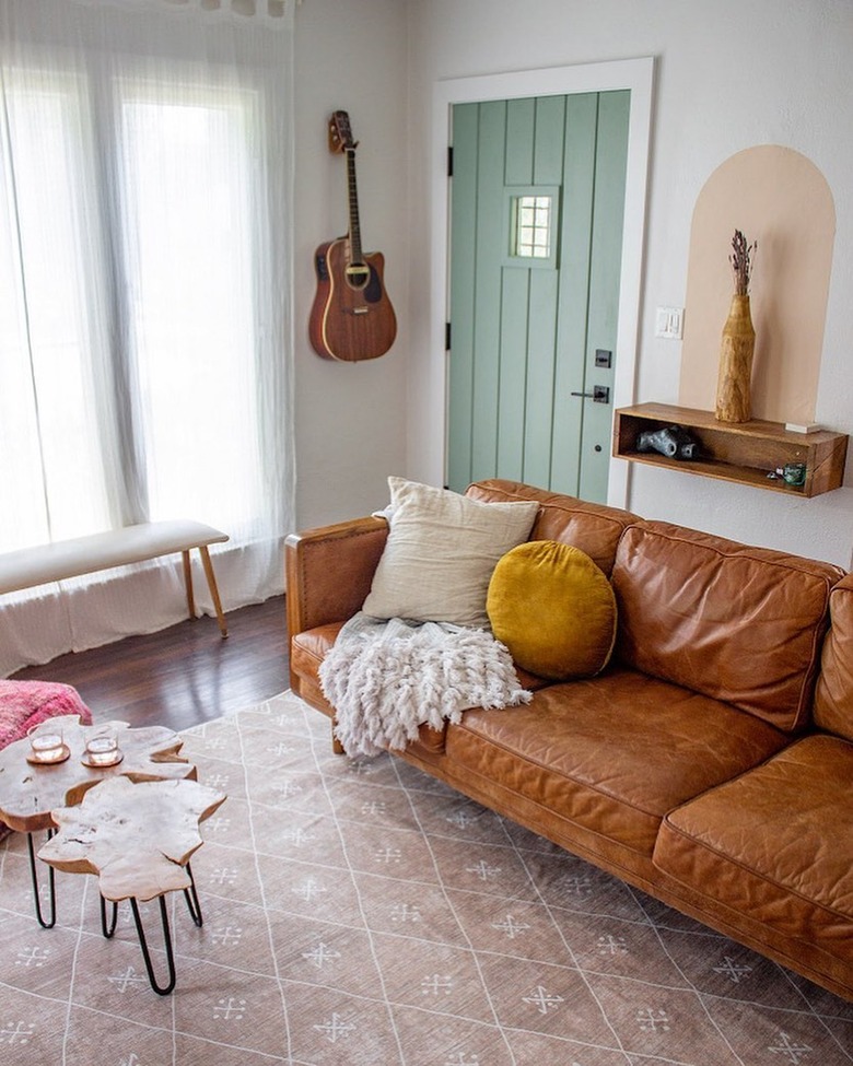 A brown leather couch styled with throw pillows and a blanket on a patterned light pink rug. A shelf and guitar hangs on the wall
