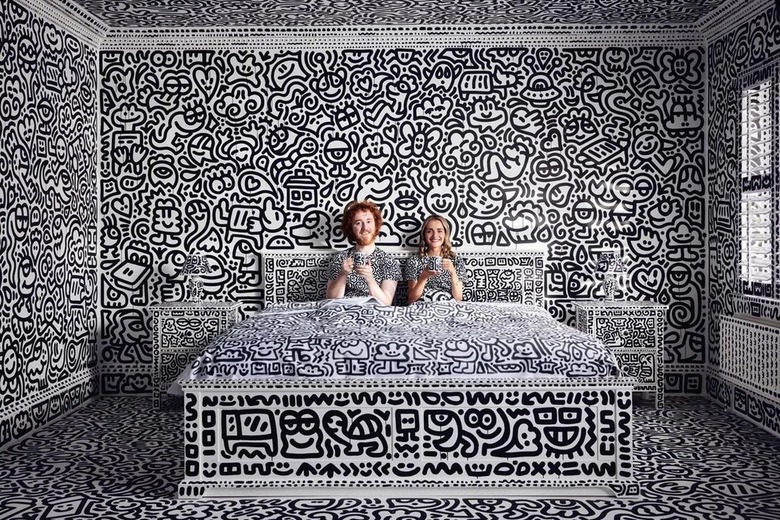 Sam Cox and his wife sitting on a doodled white bed in a completely doodled bedroom.