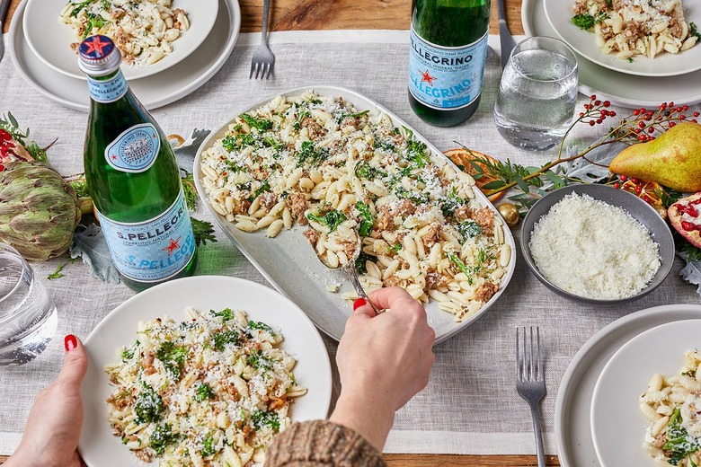 A holiday table is set with Gnocchetti con Salsiccia e Broccolini at the center of it. There is a white hand with red nail polish scooping pasta onto an individual plate. Two bottles of San Pellegrino rest on the table