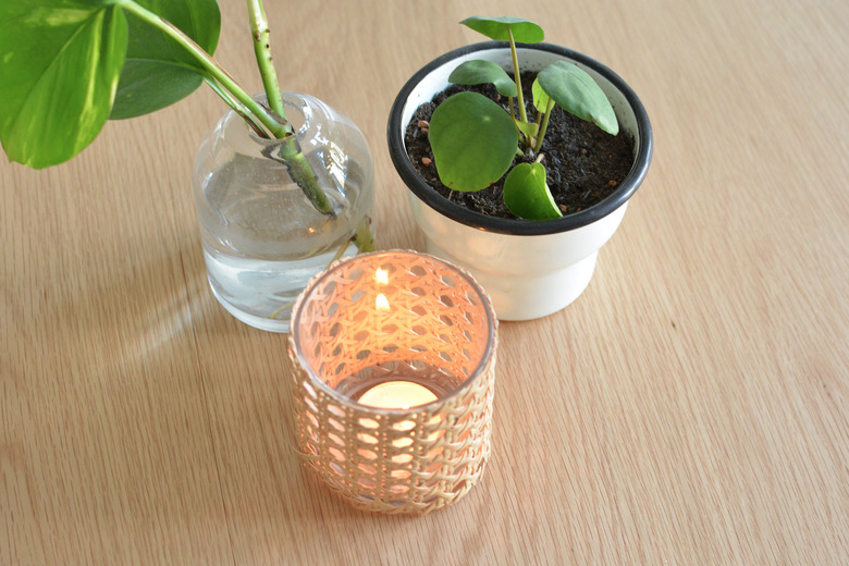 Cane candle holder with lit tealight candle inside. Next to plant pot and a glass vase with plant inside.