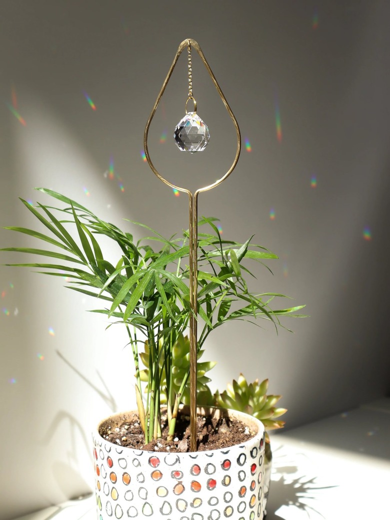 A gold wire plant stake topped with a teardrop shape that has a clear crystal hanging down the center and rainbow prisms reflected on the wall behind it.