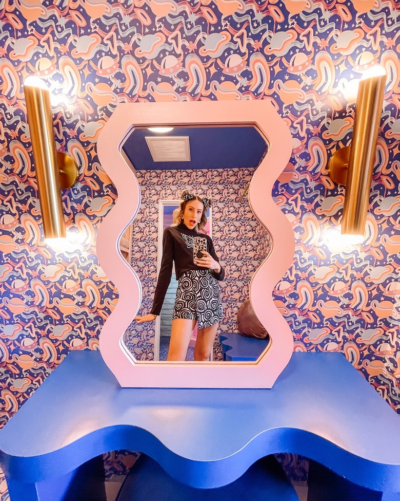 A vanity in the Trixie Motel featuring a wavy mirror lined in pink on a rich blue wavy table. In the background