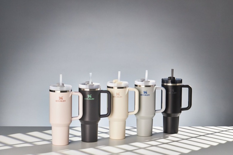 A row of five neutral colored Stanley 40oz Quencher Tumblers rest on a gray surface with a gray wall behind it. The color of the tumblers are as follows: Rose Quartz