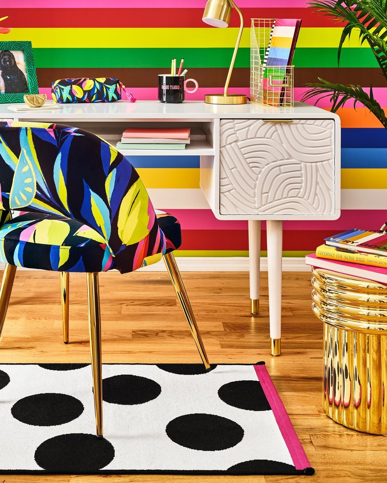 Black accent chair with bright colors resembling leaves. It sits in front of a white desk with a rainbow background on a wooden floor.