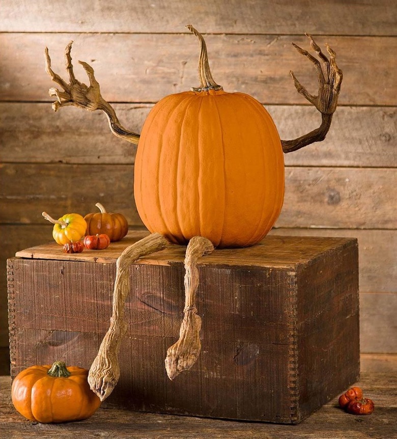 An orange pumpkin sculpture with arms and legs that look like branches. It is sitting on a wood block with small pumpkins around it.