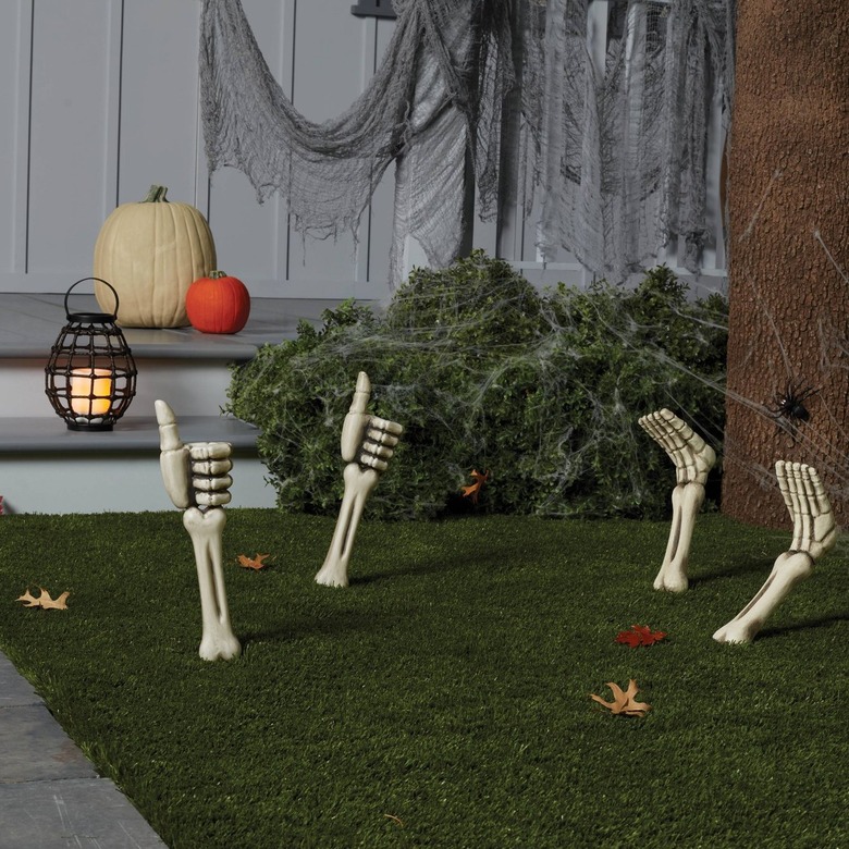 A front yard with dark green fake grass and four fake autumn-colored leaves. There are skeleton feet and hands sticking out of the ground