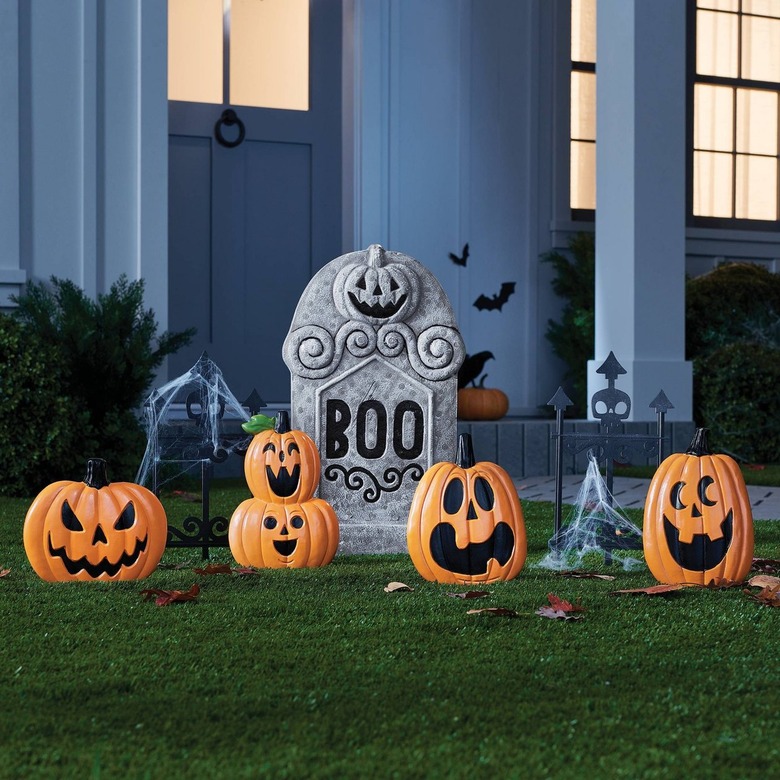 A front yard with a plastic gravestone that shows a pumpkin and says BOO. There are also four plastic jack-o'-lantern decals stuck in the green grass. Around them are fake black skull fencing draped in spider webs.