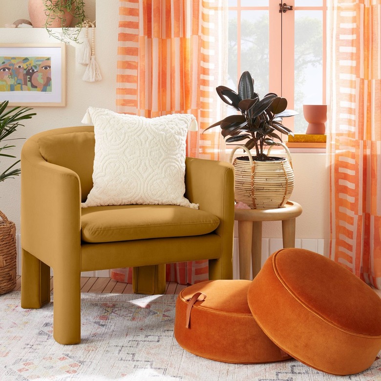 A mustard armchair with a curbed back in a living room by a window. There are two orange floor cushions on the floor