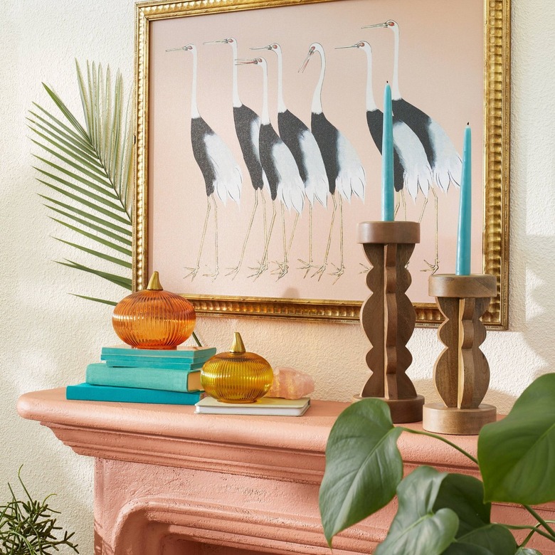 A light pink fireplace topped with tall and small curvy wood candleholders. There are also two glasses pumpkins in orange and yellow