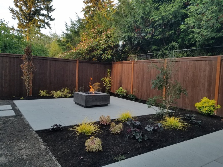 A simple concrete patio has a square fire pit and is next to a garden filled with plants and mulch.