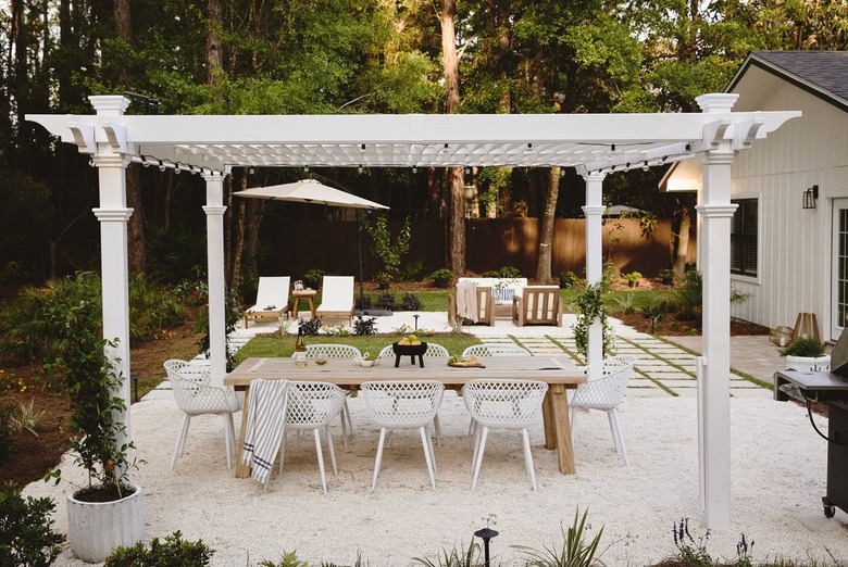 A large white pergola on white gravel. There is a large wooden dining table underneath