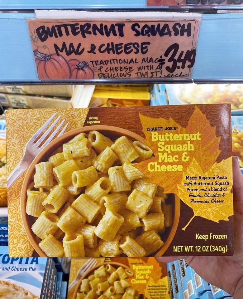 A box of Trader Joe's Butternut Squash Mac and Cheese is being held over the freezer section of a Trader Joe's aisle. The box has an orange bowl of rigatoni pasta