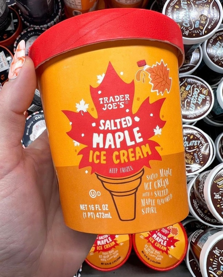 A pint of Trader Joe's Salted Maple Ice Cream is being held over the ice cream section of the freezer aisle in Trader Joe's. The carton is orange with a red top