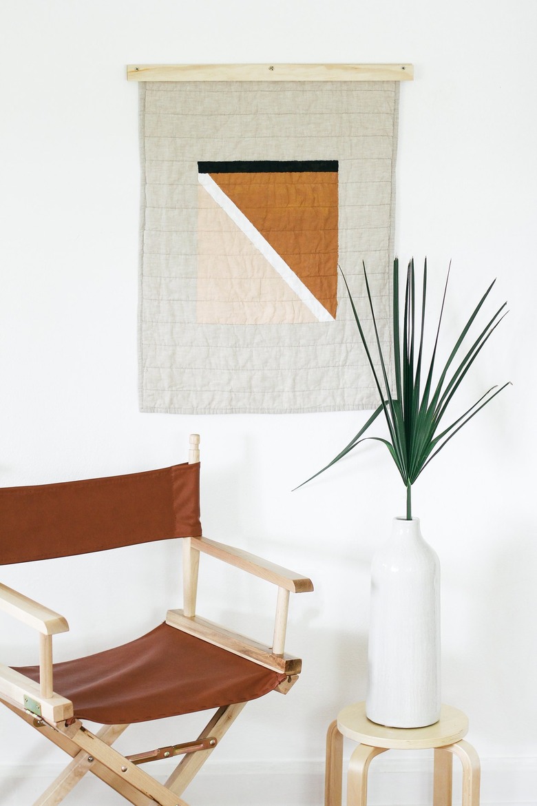 Painted quilt hanging with foldable brown chair and plant with vase on stool