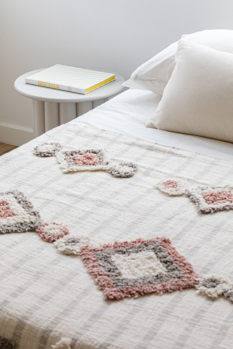 White bedding with red and purple quilting next to round white side table with book against white wall and wood flooring