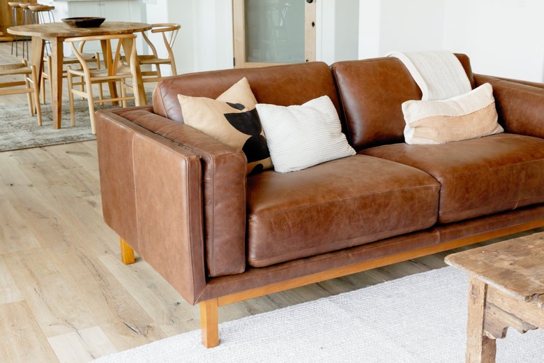 A brown leather sofa with three pillows and a white throw blanket. The sofa is on a white rug