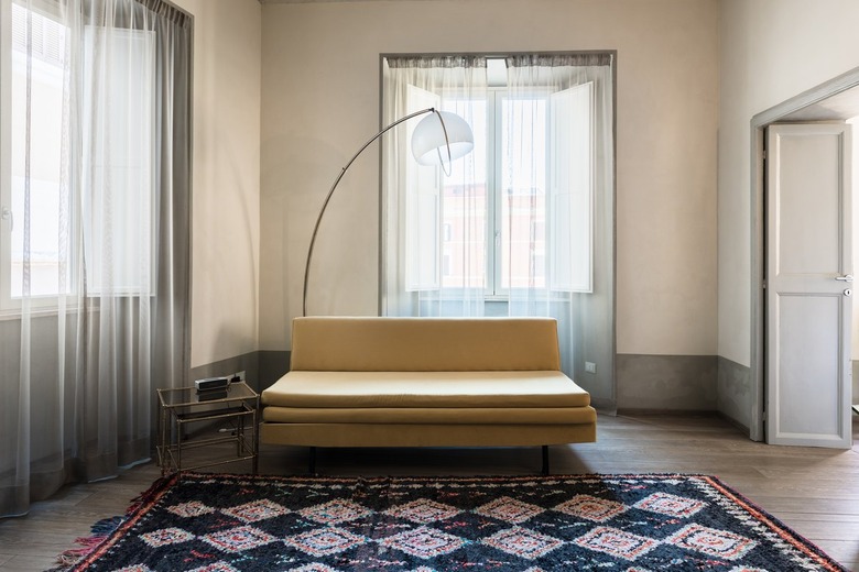 Cream-colored bench with tall lamp with small side table and rug against light tan wall with tall windows