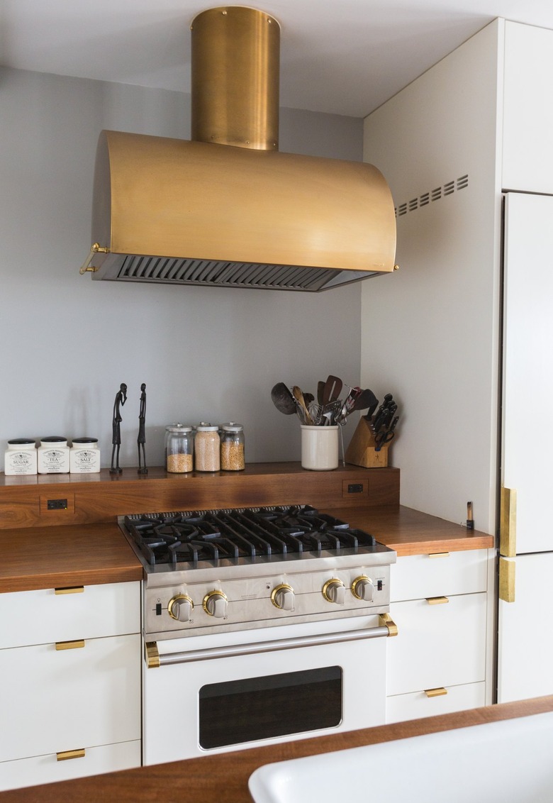 Brass stove hood over a stovetop. White cabinets with a wood counter and brass handles. Containers