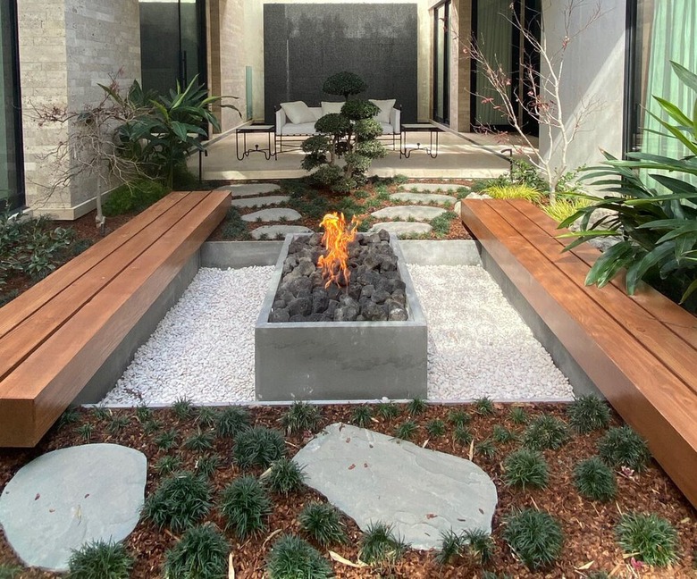 A garden with two wooden benches and a gravel patio with a concrete fire pit on top