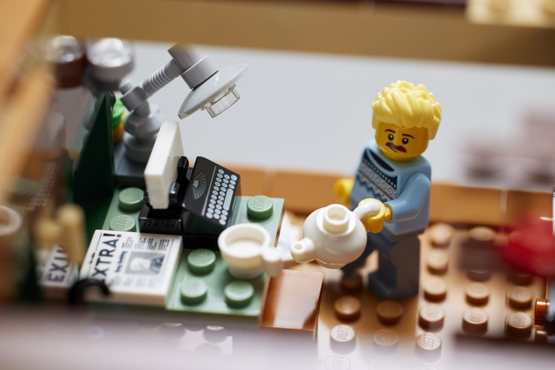A LEGO minifigure with blonde hair and blue sweater pouring coffee into a white mug on a green desk with a black typewriter.