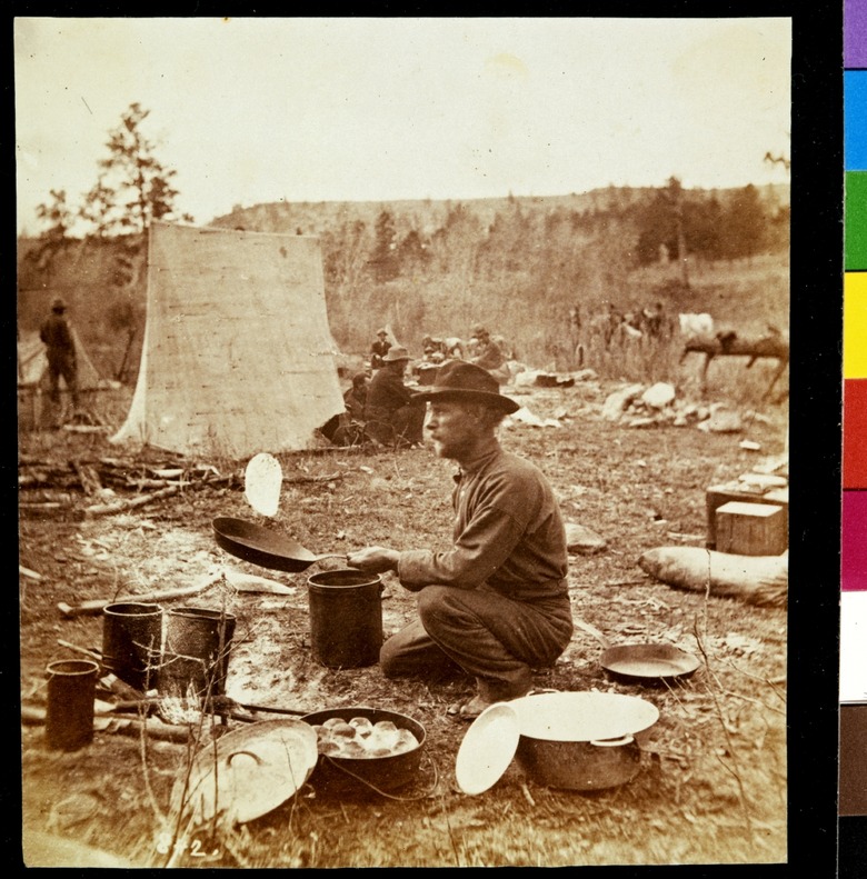 A man cooking flapjacks in a frying pan next to a Dutch oven outside