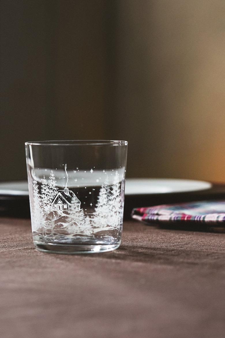 A clear glass tumbler with a pine tree and gingerbread house embossed white scene on it.