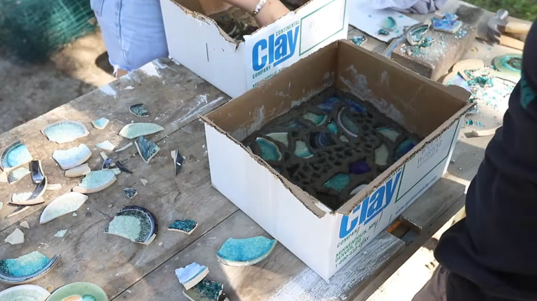 Two people arrange broken plate shards into cardboard boxes filled with concrete
