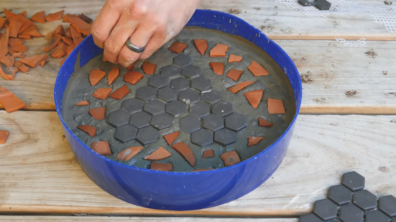 A person places small black tiles and terracotta chips into a blue bucket filled with concrete