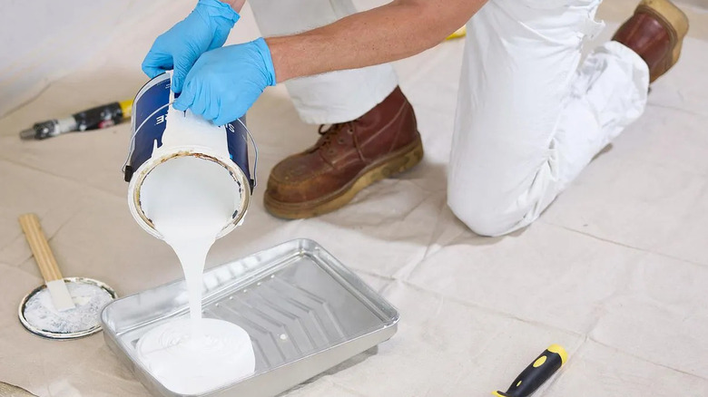 a man kneels on a drop cloth while pouring white paint into a roller pan