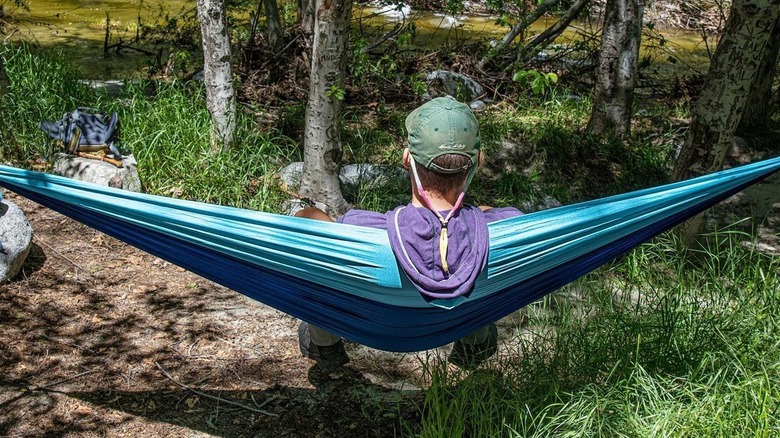 a man sits in a teal parachute hammock facing a river