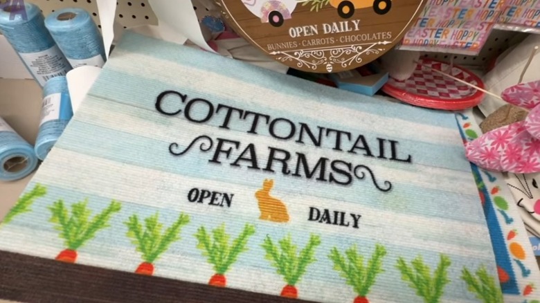 A cute spring-themed doormat on a Dollar Tree shelf, with a row of carrots, a bunny silhouette, and the words "Cottontail Farms Open Daily."