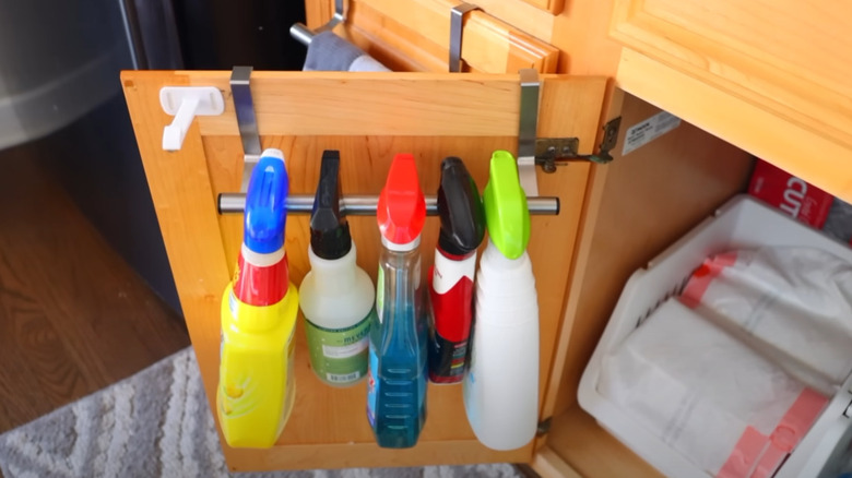 Cleaning chemical bottles hung on a dish towel rack on the inside of a cabinet door.