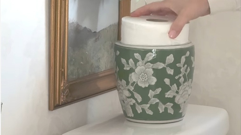 A person hiding a toilet paper roll in an ornate flower vase sitting on the back of a toilet