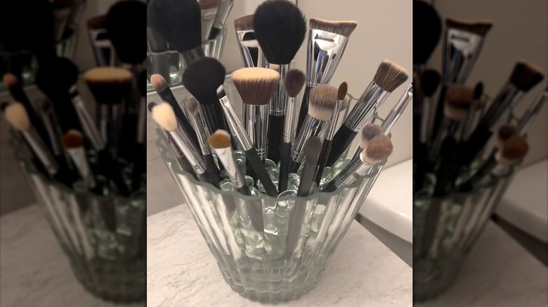 Assorted cosmetic makeup brushes sticking out of glass vase with decorative stones