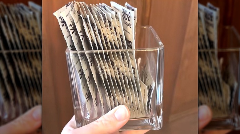 A hand holding up single-use, individually-wrapped retainer cleaning tablets in square glass vase