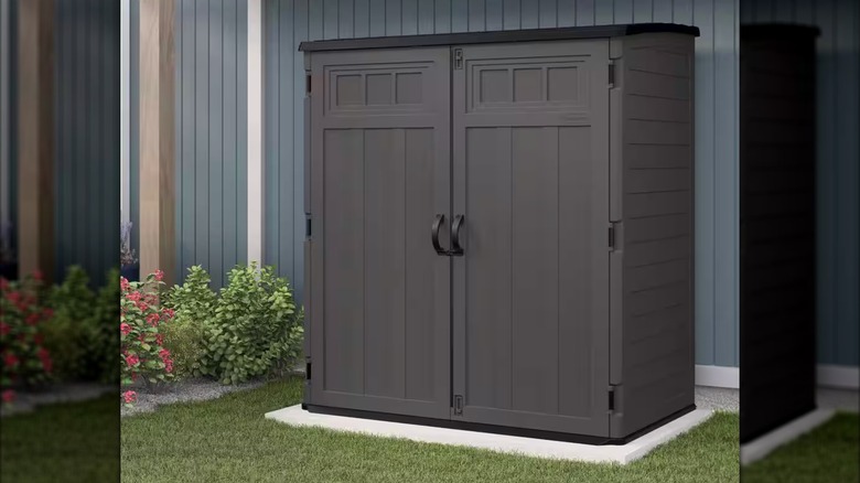 A peppercorn-colored Suncast Plastic Resin Vertical Storage Shed sits on a concrete patio next to a blue house
