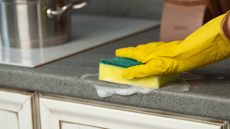 A gloved hand wiping down kitchen counter with soapy sponge