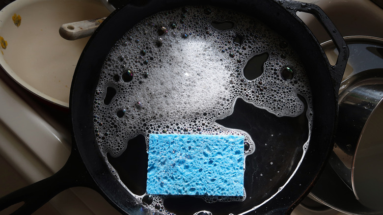 A dirty cast iron pan in the sink with soap water and a blue sponge
