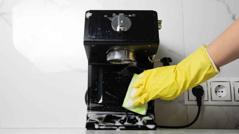 Gloved hands cleaning the exterior of a coffee machine with a soapy sponge