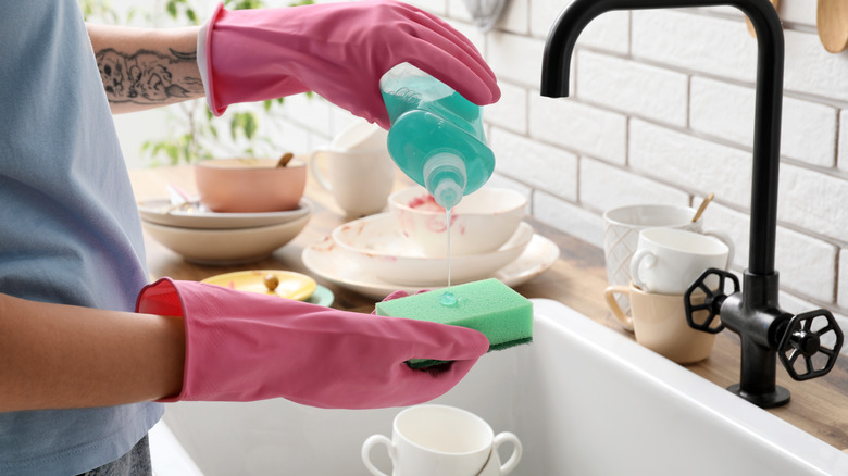 Person wearing rubber gloves pouring dish soap on a sponge over kitchen sink