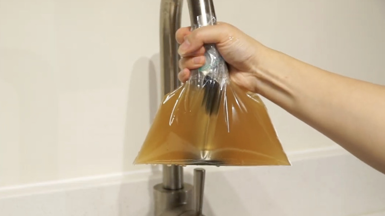 A hand holds a bag filled with apple cider vinegar over kitchen faucet end.