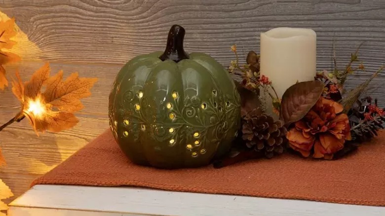 A dark green ceramic pumpkin decoration sits next to faux flowers and a cream candle