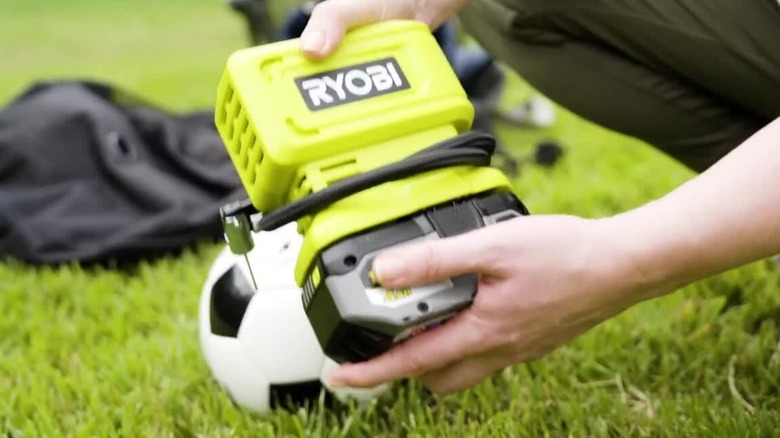 Person attaching a Ryobi battery to a Ryobi inflator with a soccer ball in the background