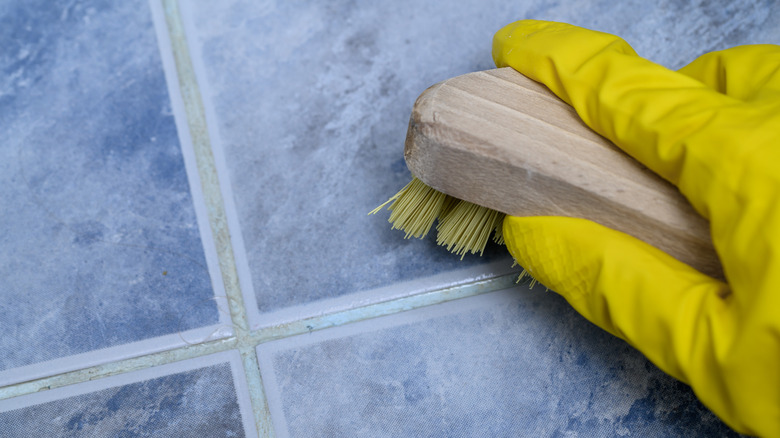 Gloved hand scrubbing blue tile and dirty grout with a brush