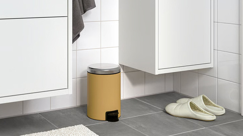 A small mustard yellow garbage can sits on a gray bathroom floor between two white floating cabinets