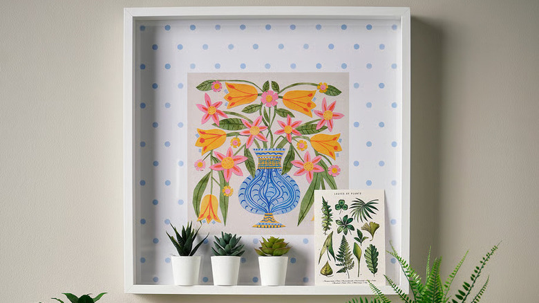 Three small plants and a small postcard sit inside the frame of a blue and white floral print