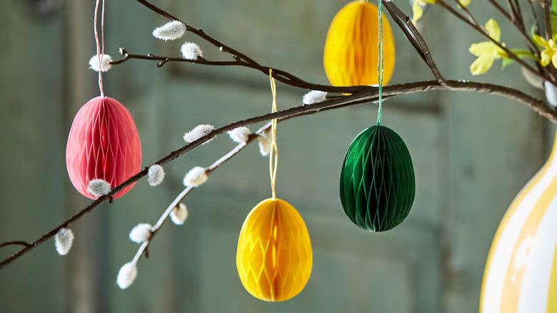 Small, decorative paper eggs hang from a thin branch in a vase